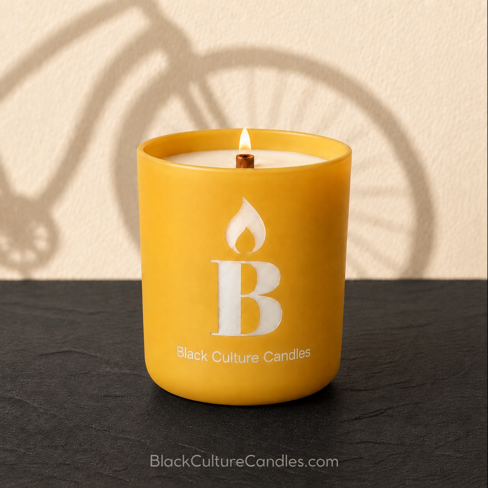 A golden yellow candle in a matte jar with an etched flame-and-letter logo and the text 'Black Culture Candles' on the front, lit with a small flame, set on a dark surface against a beige wall with a bicycle shadow.