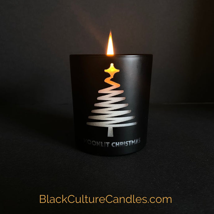 Moonlit Christmas is a limited edition candle in a matte black jar with a modern pine tree engraving. The candle is partially burned, revealing a warm and inviting glow, casting a cozy atmosphere. The label on the jar prominently displays the Black Culture Candles logo. BlackCultureCandles.com