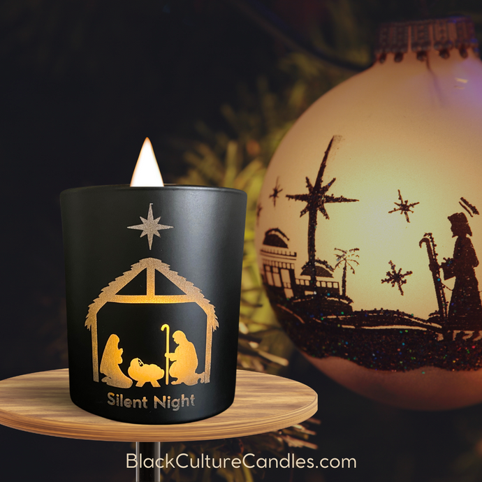 A matte black candle etched with a delicate nativity scene, glowing softly as its flam flickers. The crackling wood wick adds warmth, surrounded by holiday decor.