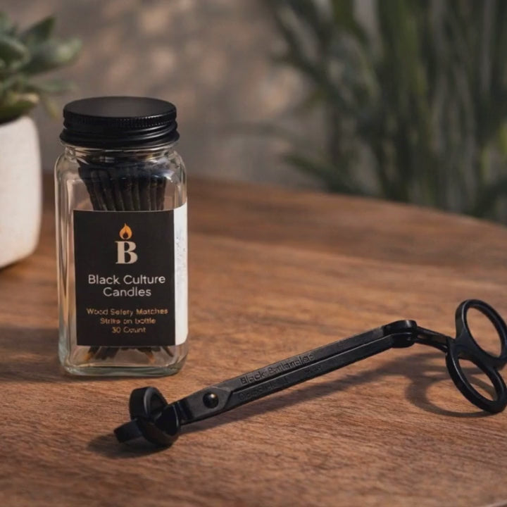 Black Culture Candles candle accessory set with wood safety matches in a glass jar and black metal wick trimmer on a wooden table.